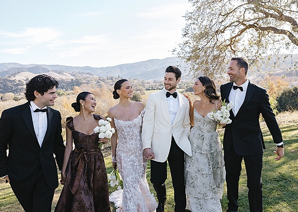 Wedding party portrait of bride and groom with friends walking arm in arm, bride in strapless lace dress, mountains and blue sky behind