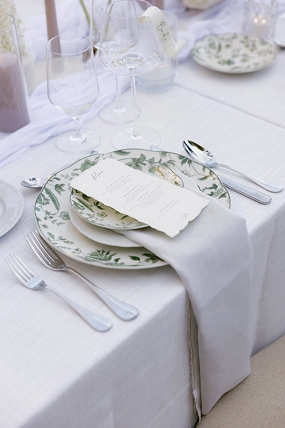 Reception tablescape with wedding place setting, green floral rim plates, deckled menu card, gray napkin, silver flatware, and taper candles on linen runner