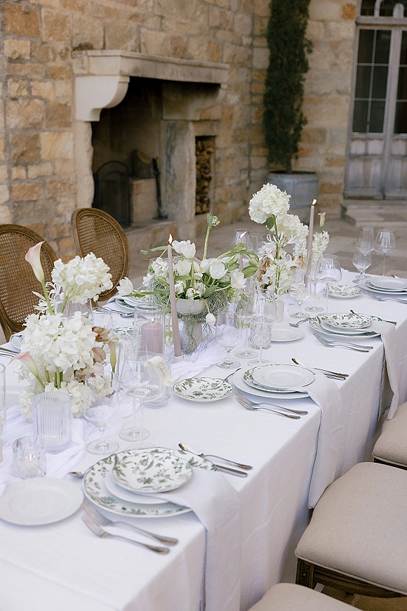 Reception tablescape with white tablecloth and hydrangea centerpieces, pastel taper candles, and floral plates on a patio by a stone wall fireplace