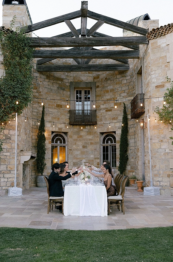 Reception tablescape with outdoor banquet table set in a stone courtyard, taper candle accents, floral centerpiece, and string lights overhead