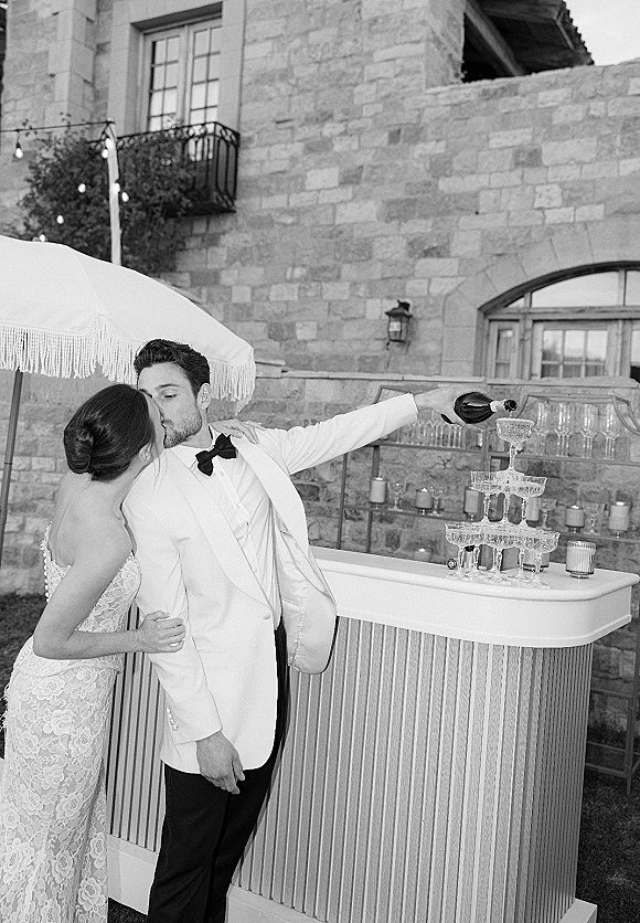 Wedding kiss portrait of bride in a strapless lace dress and groom in a white tuxedo beside a champagne tower on a stone patio with string lights