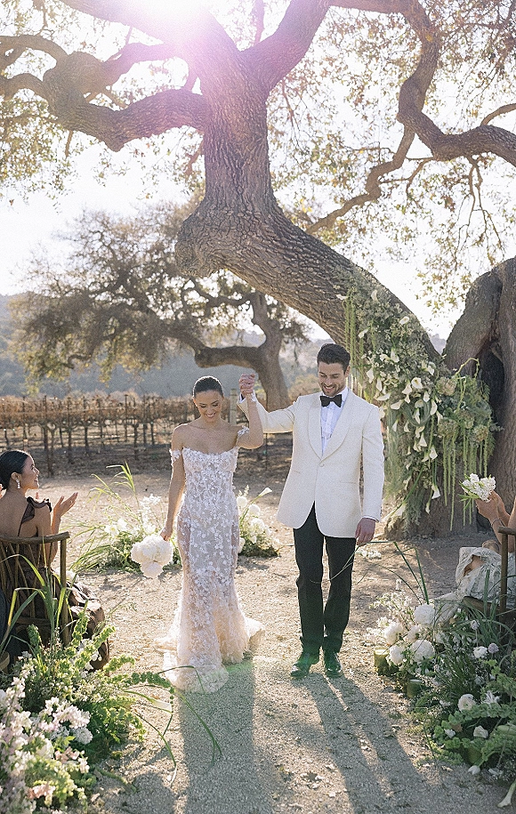 Wedding recessional as bride and groom walking aisle hand in hand, bride with white bouquet, groom in white tuxedo, sunlit vineyard under tree