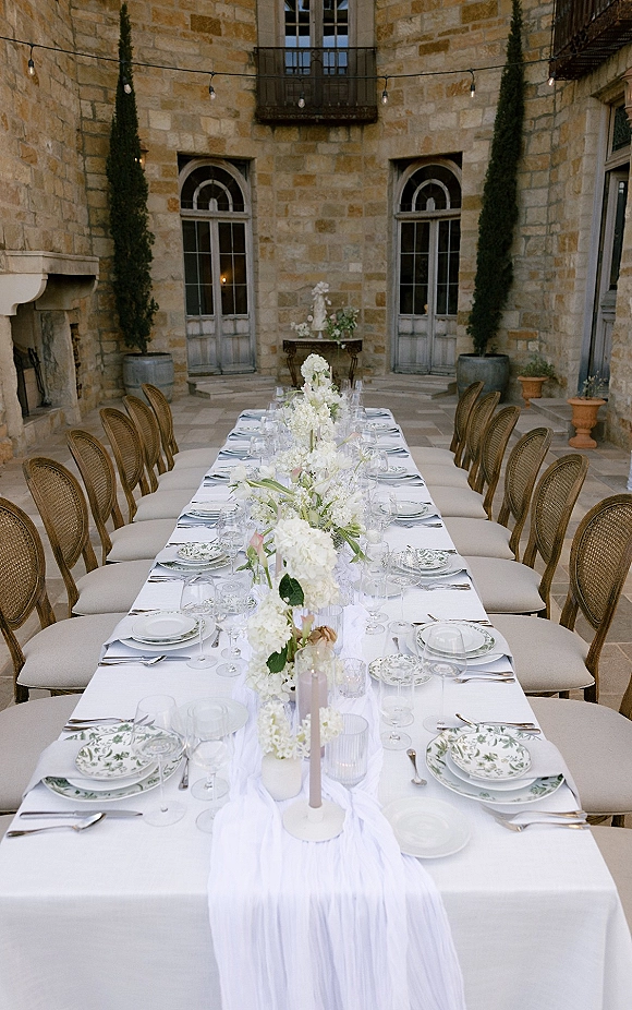 Reception tablescape with long banquet wedding table, white gauze runner, floral centerpiece, taper candles and glassware in a stone courtyard under string lights