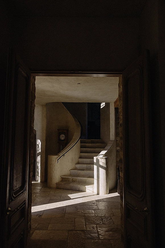 Wedding venue interior with a stone staircase and curved rail, antique clock and wooden doors, lit by window light on tiled floors