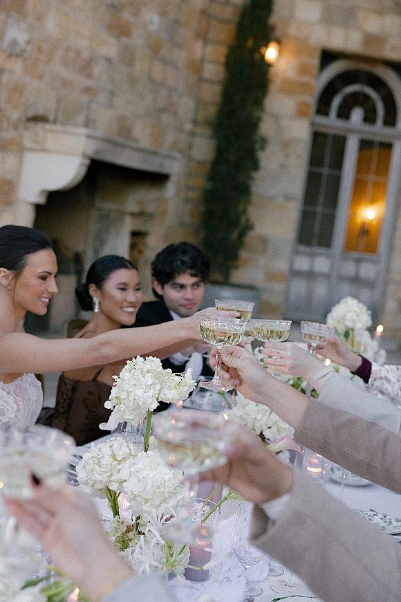 Wedding toast as bride and groom clink champagne coupe glasses over a candlelit table with white florals on a stone courtyard patio
