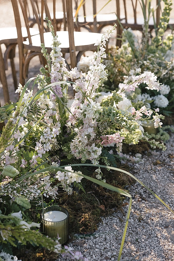 Ceremony aisle florals with pastel blooms and greenery on a gravel walkway, dotted with moss and glass votive candles beside wooden chairs