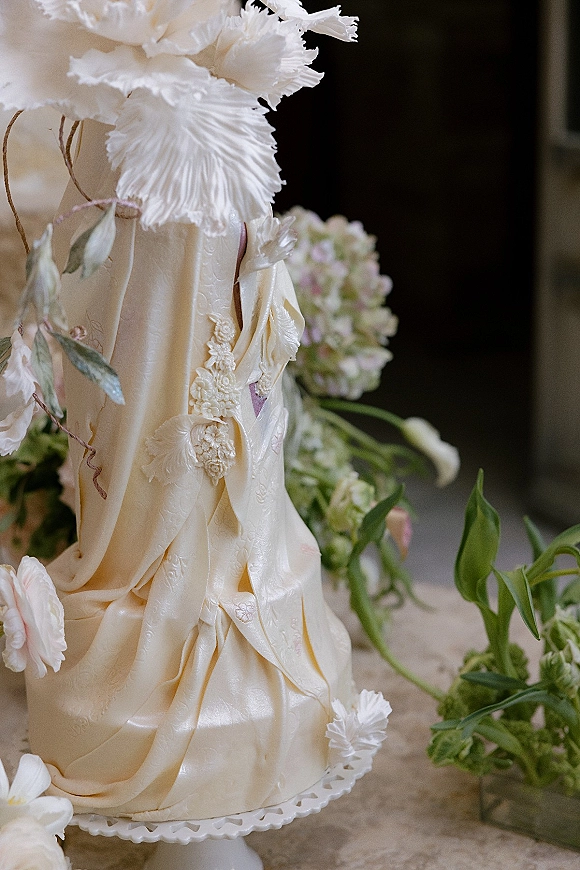 Wedding cake with buttercream draping and lace piping, topped with sugar flowers and greenery on a cake stand against a dark interior background