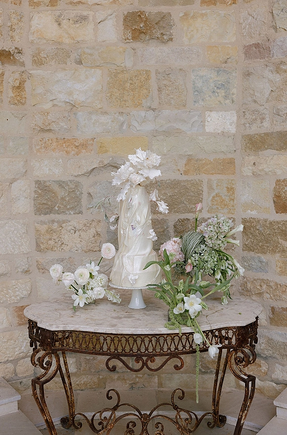Wedding cake display featuring a white buttercream wedding cake on a stand with fresh flowers and greenery on marble, set against a stone wall