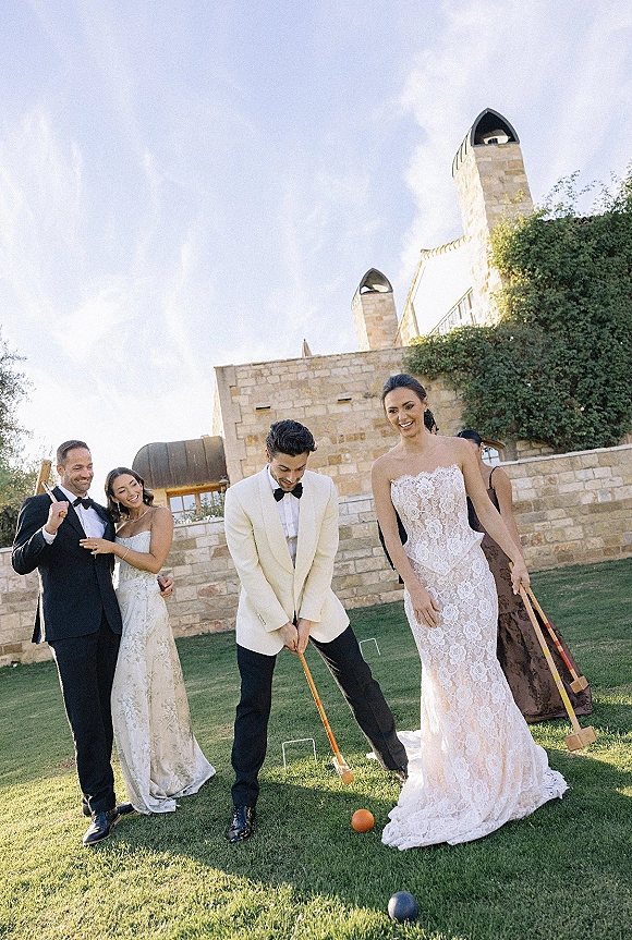 Wedding lawn games as bride and groom play wedding croquet in tuxedo and lace dress on a grassy lawn by a stone venue under blue sky