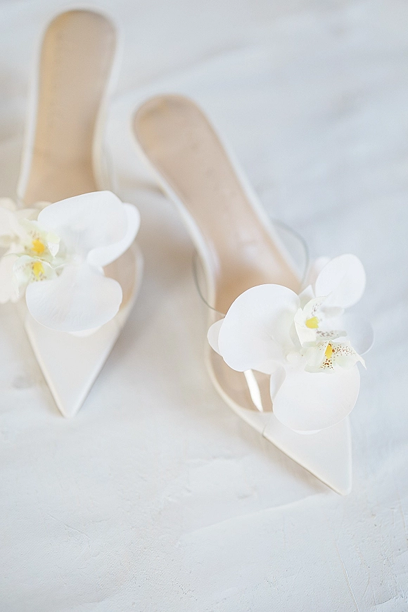 Bridal shoes, white pointed-toe heels with orchid shoe clips and floral appliques, styled on a white textured surface flat lay