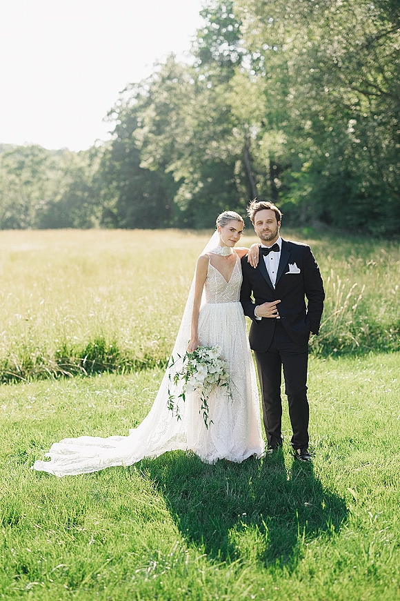 Couple portrait of bride and groom in a sunlit meadow, her veil and train flowing as they hold a white greenery bouquet
