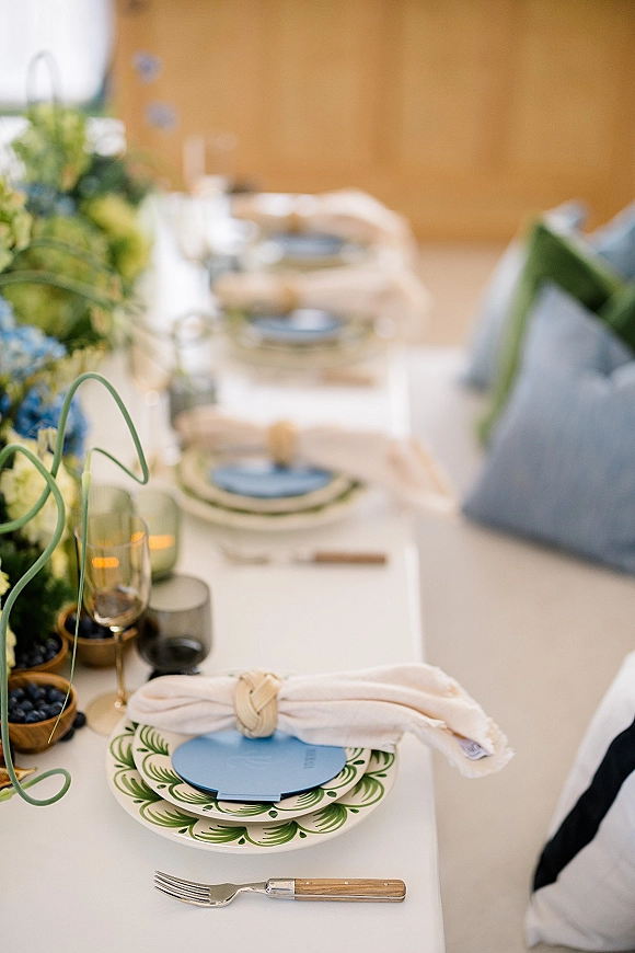 Reception tablescape on a long banquet table setup with floral centerpiece, green foliage and blue flowers, taper candles, colored glassware, and place cards