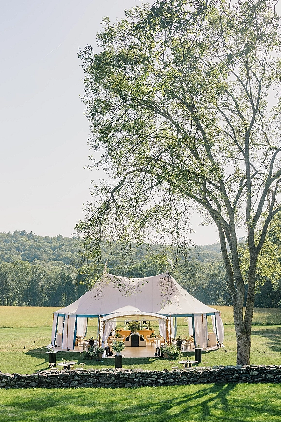 Outdoor reception tent with white draping, lounge seating and bar, styled with floral arrangements and lanterns in a grassy field by a stone wall