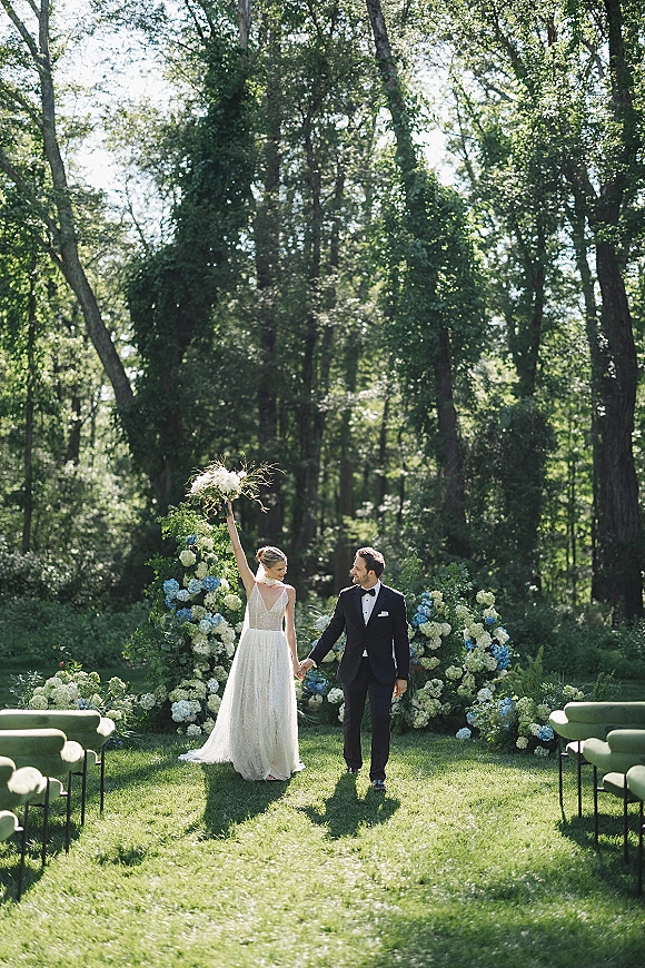 Recessional moment as bride and groom walk the aisle holding hands, bride lifting bouquet overhead in sunlit forest ceremony setting