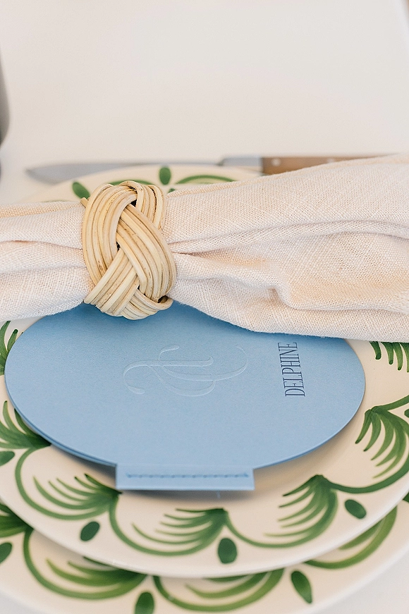 Wedding place setting with a woven napkin ring, linen napkin, blue menu card on a green patterned plate and flatware on a white tabletop