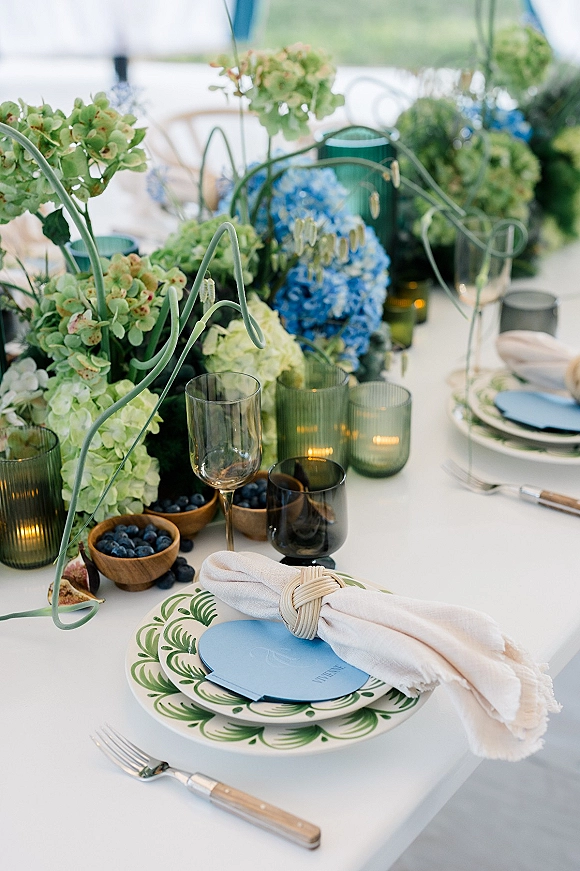 Reception tablescape with blue and green hydrangea centerpiece, taper candles, green glass goblets, and blueberries on a white tented table