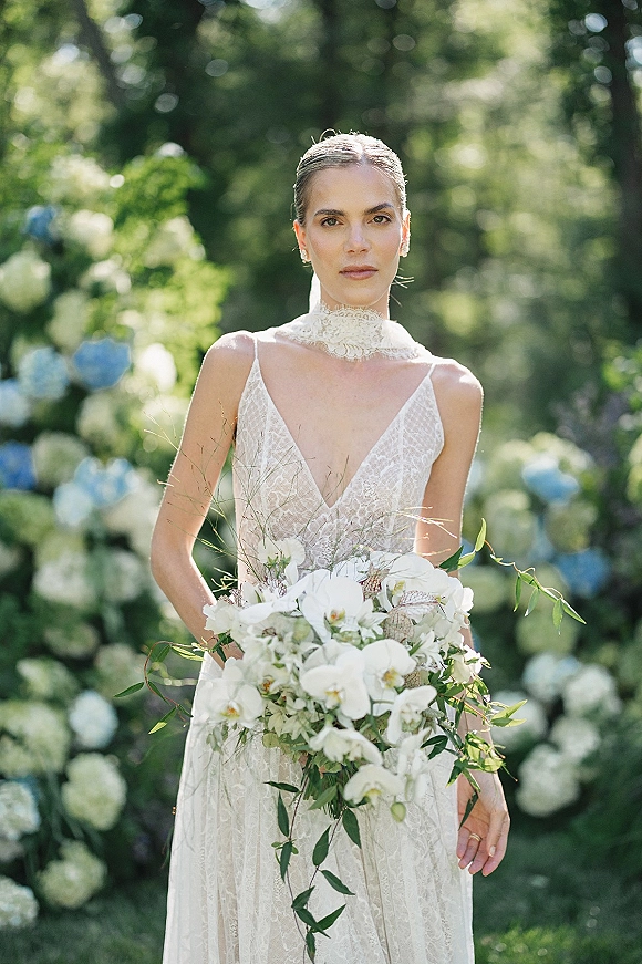 Bridal portrait of a bride holding a white orchid bouquet, wearing a high neck lace wedding dress in a lush garden with hydrangeas