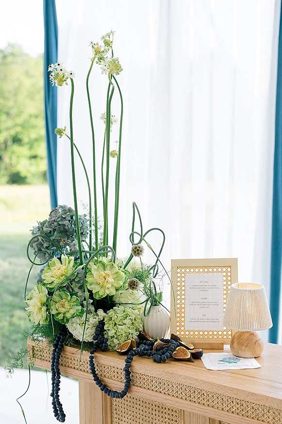 Wedding welcome table with framed wedding welcome sign, tall hydrangeas and greenery, beaded garland and figs by sunlit window curtains