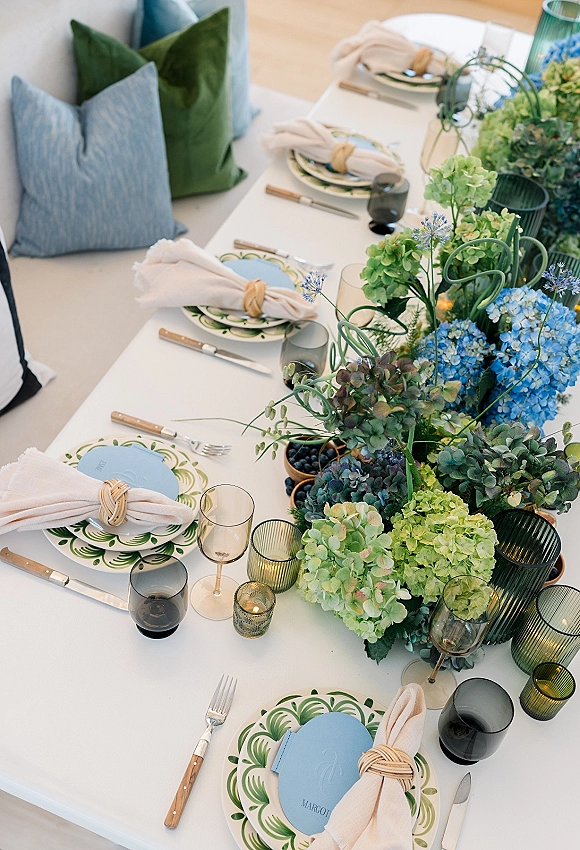 Reception tablescape with a hydrangea wedding centerpiece, blue place cards, green votives, candles, blueberries, and white banquette seating