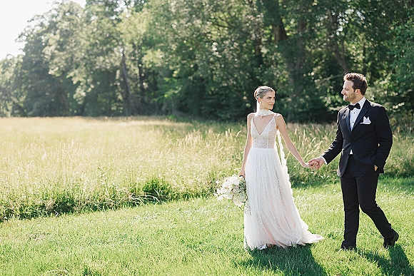 Couple portrait of bride and groom holding hands, bride with bouquet and lace dress, walking through sunlit meadow grass with trees