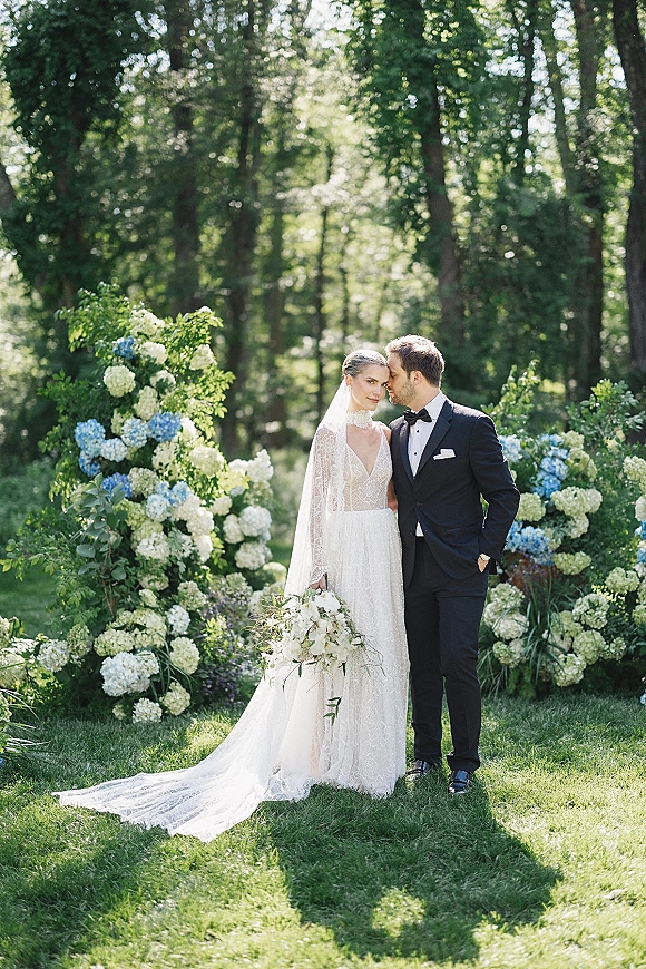 Couple portrait of bride in lace gown with cathedral veil leaning on groom in tuxedo, holding hydrangea bouquet amid woodland trees