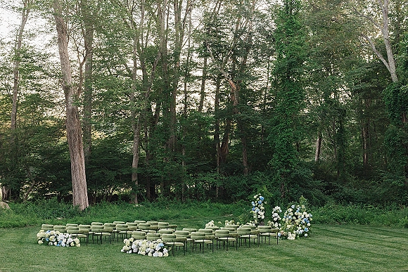 Ceremony seating setup with green upholstered chairs in rows, surrounded by ground hydrangea and white-blue florals on a forest lawn