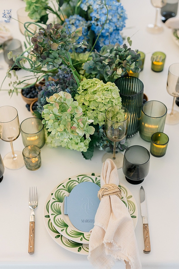 Reception tablescape with hydrangea centerpiece, greenery, and votive candles beside patterned plates, menu cards, and glassware on a white tablecloth