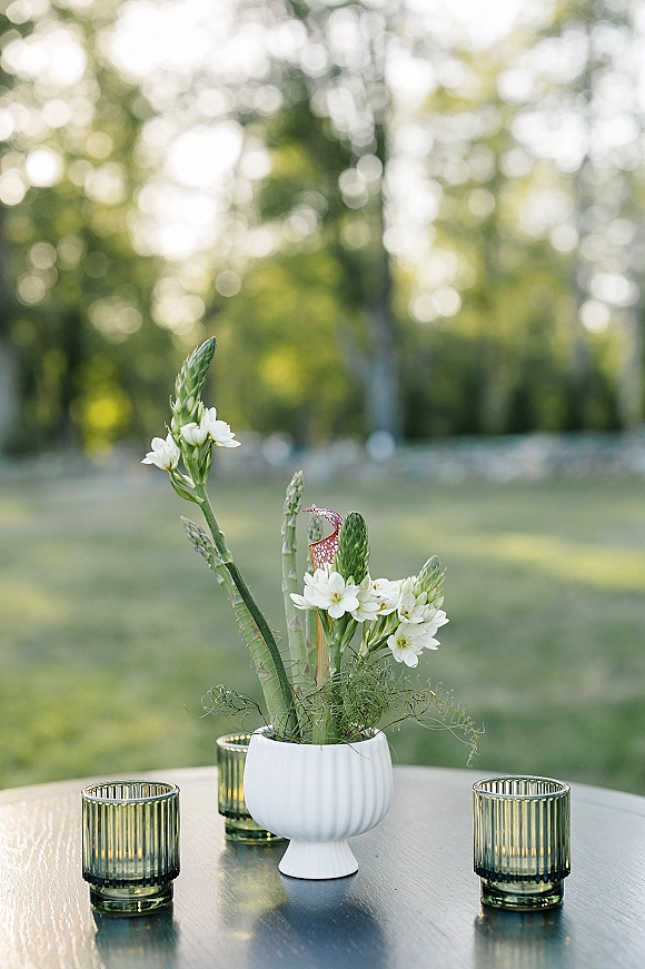 Wedding centerpiece with white floral arrangement and greenery in a white ceramic vase, paired with green glass votive candles on a lawn