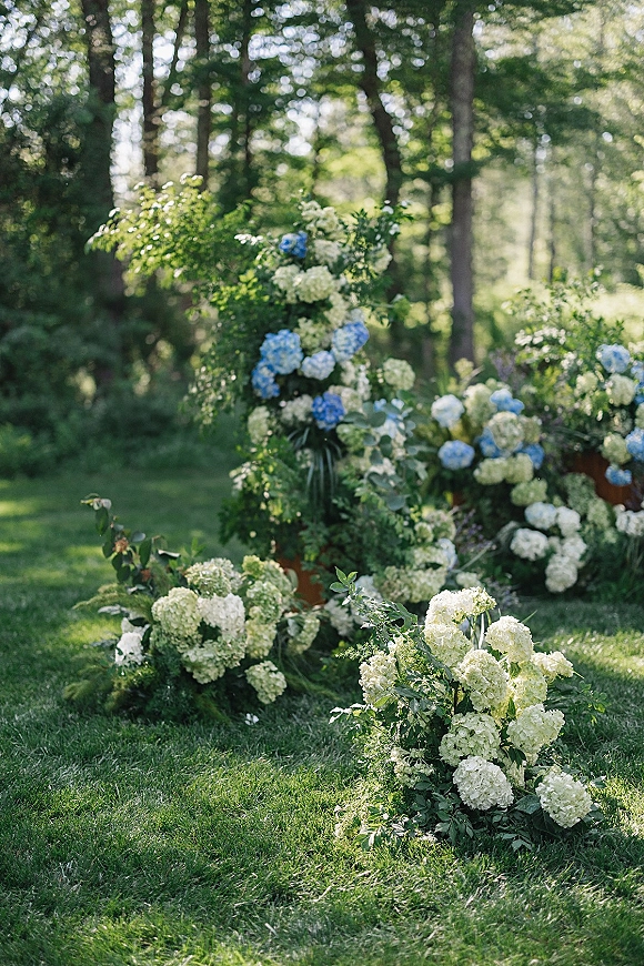 Ceremony aisle florals with hydrangea aisle flowers and greenery arranged low along the path on a garden lawn with trees in back
