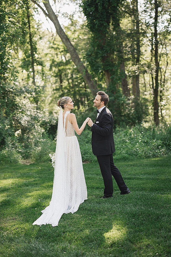 Couple portrait of bride and groom holding hands, facing each other on a sunlit garden lawn, her long veil and bouquet visible
