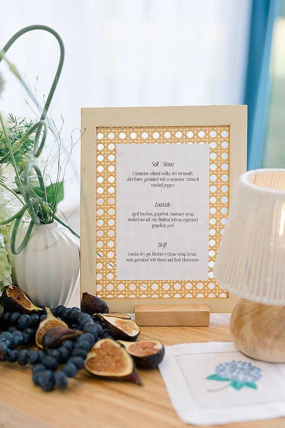 Wedding bar menu framed in a rattan cane frame with calligraphy card, florals, and figs on a wood tabletop in window light