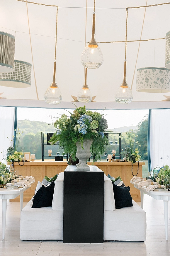 Reception tablescape with head table centerpiece in a large urn floral arrangement, pendant lights, candles, and tented windows facing hills