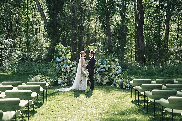 Ceremony moment as bride and groom exchange vows under a hydrangea wedding arch, veil and bouquet glowing in dappled forest light