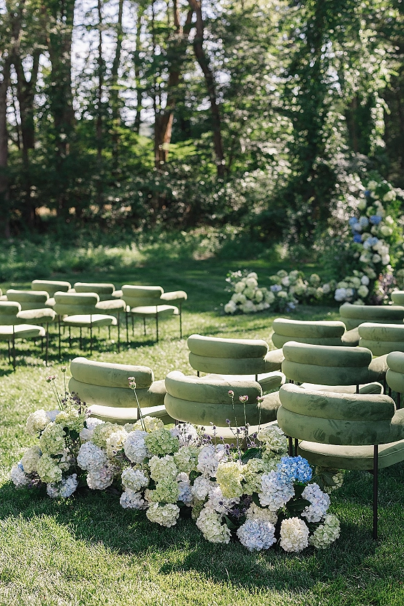 Ceremony seating with green lounge chairs arranged on a grass lawn, bordered by low hydrangea florals in a wooded garden setting