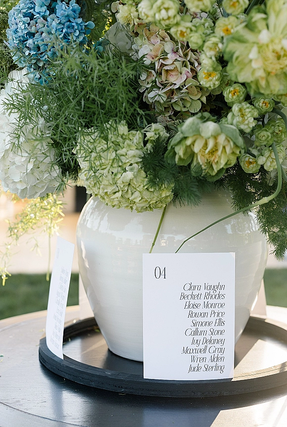 Wedding escort cards arranged on a black round tray beside a white ceramic vase of hydrangeas and greenery on a sunlit lawn table