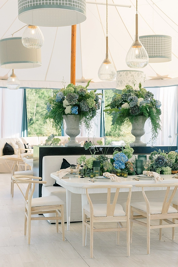 Reception tablescape with hydrangea wedding centerpiece in stone urns, teal candle holders and white place settings under a draped tent with pendant lights