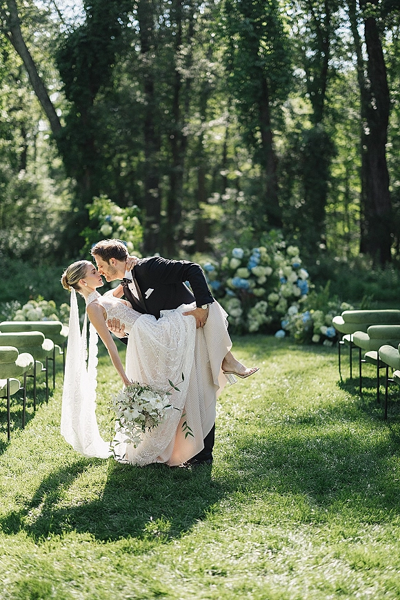 Wedding kiss portrait as groom dips the bride for a dip kiss wedding photo, veil flowing beside the ceremony aisle in a sunlit garden