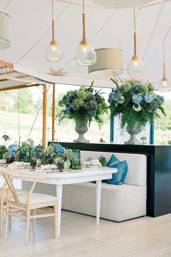 Reception tablescape with blue hydrangea centerpiece in stone urns, greenery garland, taper candles, and teal votives under a tent ceiling