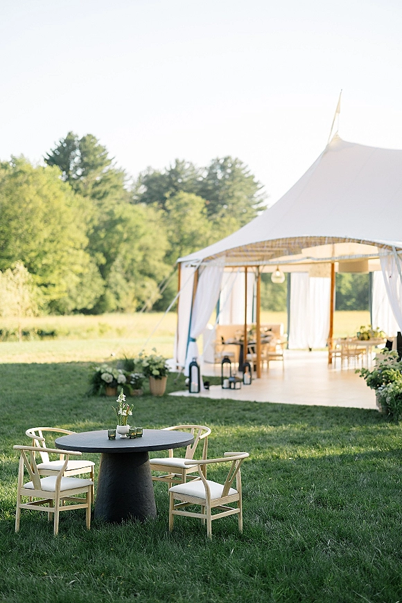 Outdoor reception tent with a wood dance floor and round cocktail table, accented by lanterns and florals on a grassy meadow lawn