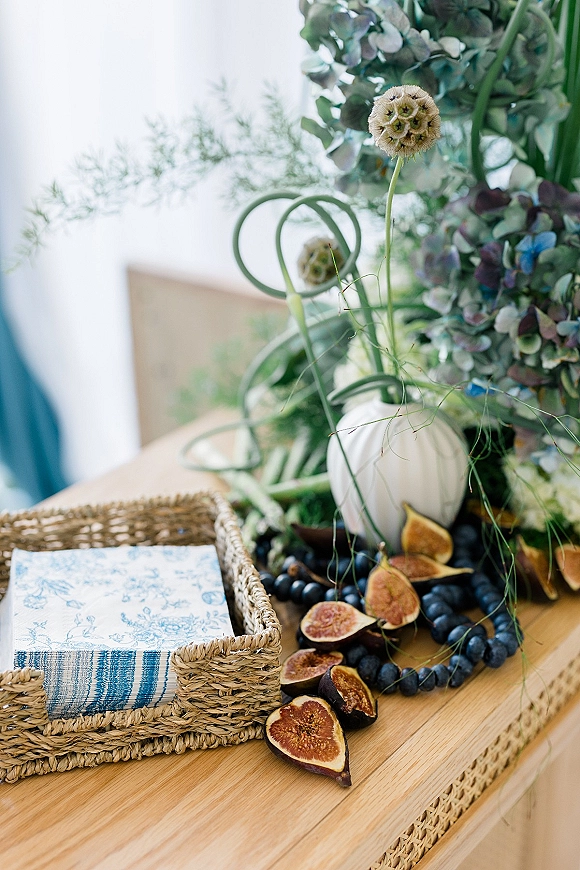 Reception tablescape with wedding fruit display of figs and grapes beside a floral arrangement, blue and white napkins, and a small pumpkin on wood table