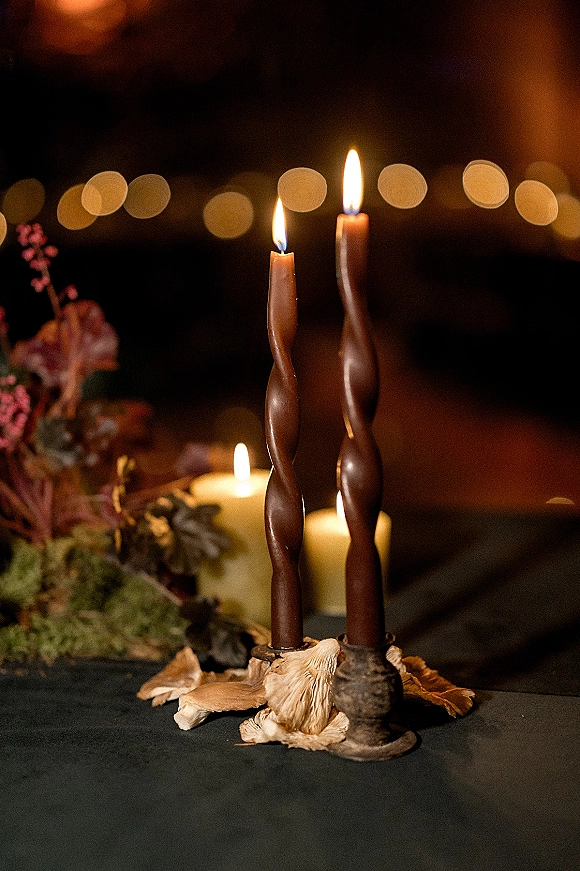 Wedding candle decor with twisted taper candles and pillar candles, dripping wax, dried flowers and greenery on a dark tablecloth in dim bokeh light