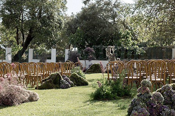 Ceremony setup with outdoor ceremony seating of bentwood chairs, moss mounds and pastel florals on a garden lawn by trees and iron fence