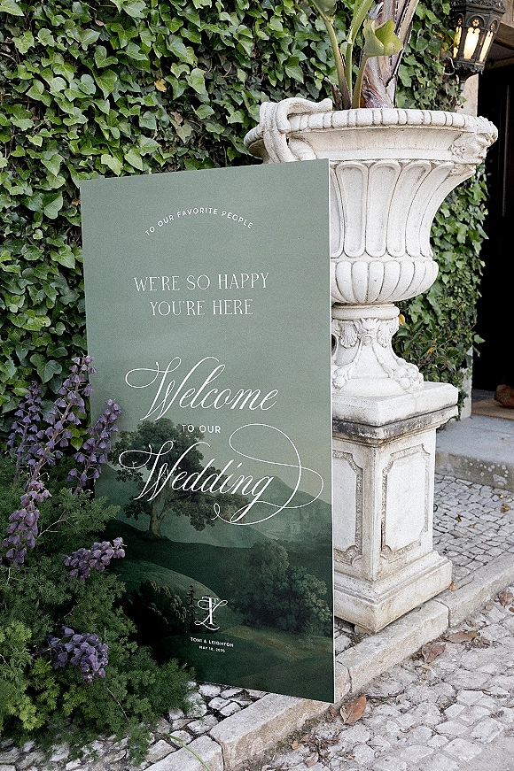Wedding welcome sign with welcome to our wedding sign calligraphy and illustrated landscape, beside a stone urn with greenery and purple flowers by an ivy wall