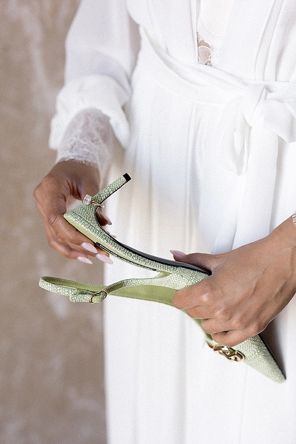 Bridal shoes in pastel green slingback heels held by the bride, showing lace robe cuffs, manicure, and engagement ring by a neutral wall