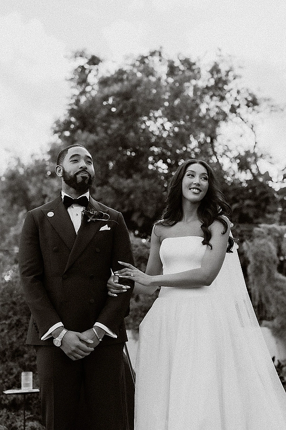 Couple portrait in a black and white wedding portrait style, bride in strapless gown with long veil holding groom’s arm in a garden setting