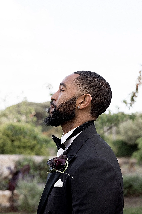 Groom portrait in side profile wearing a black tuxedo with bow tie and boutonniere, standing outdoors by trees and hillside under open sky