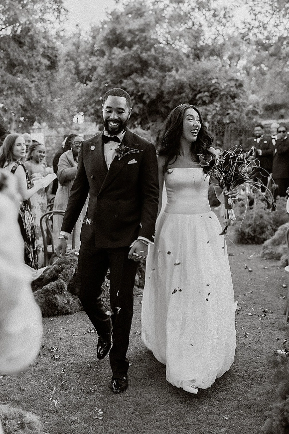 Wedding recessional as bride and groom walking aisle holding hands, bride with bouquet and tulle skirt amid petal toss on garden path