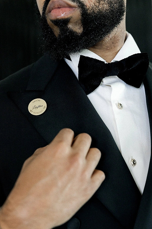 Groom portrait showing groom tuxedo details with black tuxedo jacket, bow tie, white shirt studs, lapel pin, and beard on dark background