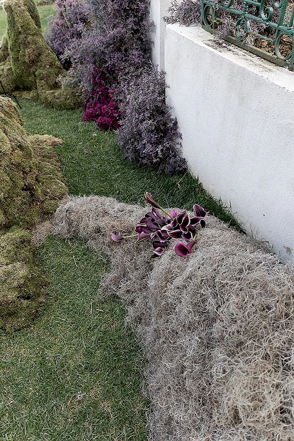 Wedding aisle decor with aisle ground florals on a moss runner, featuring purple calla lilies and lichen along a garden lawn by a white stucco wall