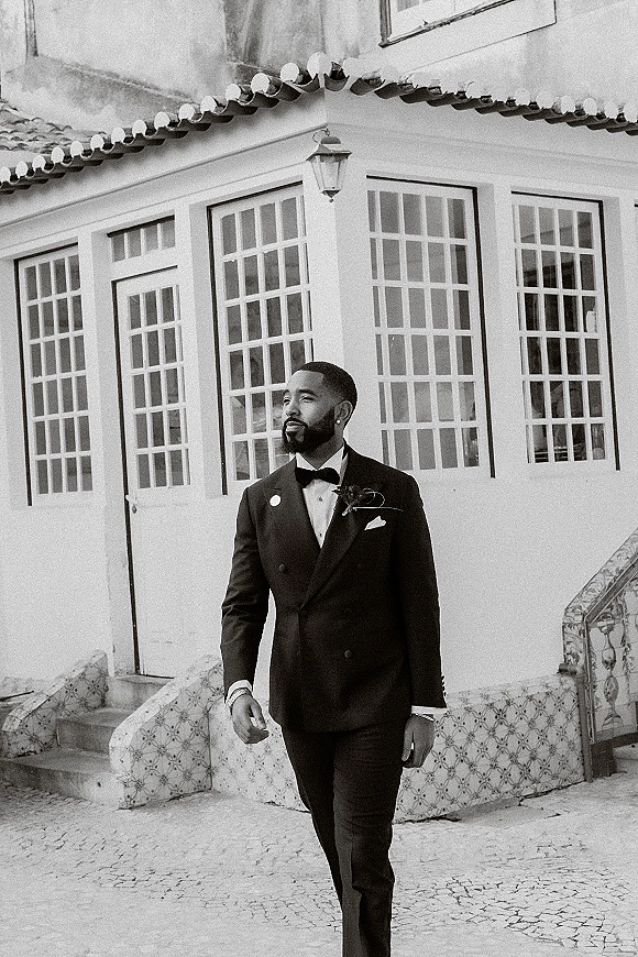 Groom portrait in a black tuxedo with bow tie and boutonniere, standing by a white villa facade with glass windows and tiled steps